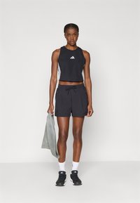 Čierny zastrihnutý tielko s bielym logom, v kombinácii s čiernymi šortkami na šnúrku a prednými vreckami. Model drží šedú osuška.