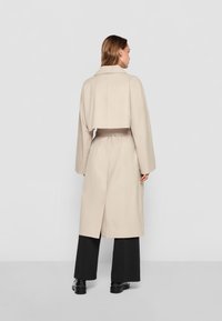 Beige trench coat with a cropped design, oversized sleeves, and a belt. It features a back vent and is styled with black wide-leg pants.