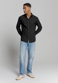Zwarte langek mouw shirt met knopen, gecombineerd met lichtblauwe jeans en beige sneakers. Model staat met handen in de zakken tegen een neutrale achtergrond.