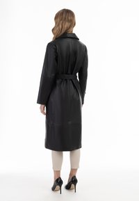 Cappotto trench in pelle nera con vita cintata, maniche lunghe e colletto classico, caratterizzato da una texture liscia e dettagli delle cuciture.