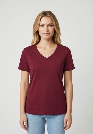 Donna bionda con occhi azzurri che indossa una T-shirt bordeaux a V semplice e jeans azzurri, in piedi davanti a uno sfondo uniforme.
