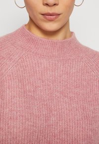 Gros plan d'une personne portant un pull en maille rose duveteuse et des boucles d'oreilles en forme d'anneau, montrant le cou et la partie inférieure du visage.