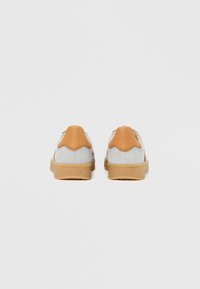 Paar hellgrauer Sneaker mit hellbraunen Leder-Fersenpolstern und Sohlen aus Gummikautschuk, von hinten vor einfachem Hintergrund betrachtet.