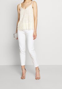 Débardeur crème à bordure en dentelle avec broderie florale délicate, associé à un pantalon blanc ajusté et des talons argentés à lanières, tenant une pochette argentée.