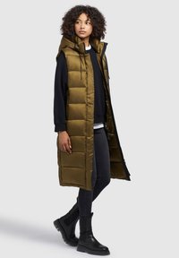 Hooded, olive green puffer vest with quilted design, front zipper, and snap closure; worn over a black sweatshirt and skinny jeans, paired with black boots.
