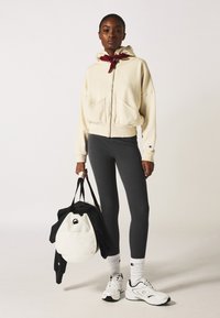 Beige rits-jas met een grote capuchon en zak, gecombineerd met donkergrijze leggings. Het model houdt een witte tas vast en draagt witte sport schoenen.
