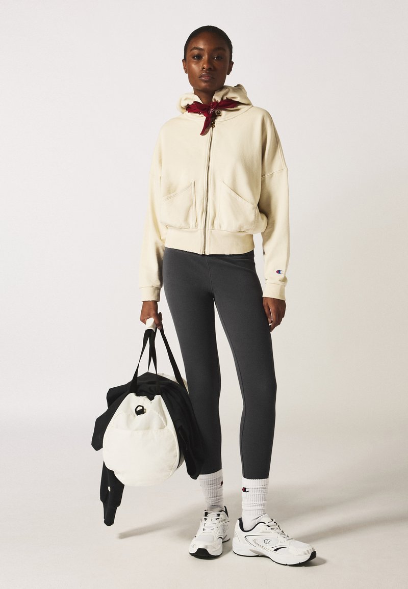 Felpa beige con zip, dotata di un ampio cappuccio e tasca, abbinata a leggings grigi scuri. Il modello tiene una borsa bianca e indossa scarpe da ginnastica bianche.