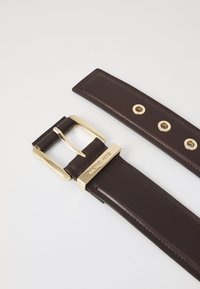 Bruine leren riem met een gouden metalen gesp. Beschikt over meerdere gaatjes voor verstelling en heeft een gladde textuur. Opvallende merkaanduiding op de gesp.