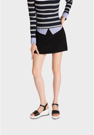 Donna con minigonna nera, top a maniche lunghe a righe e sandali con zeppa bianchi e neri, che posa con una mano sul fianco.