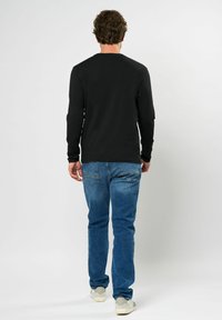 Schwarzes Langarmshirt, kombiniert mit blauen Jeans und weißen Schuhen, von hinten betrachtet; lässiger Schnitt und glatte Textur sichtbar.