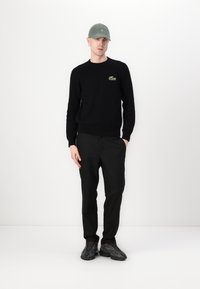 Pull en tricot noir avec logo vert, pantalon noir et casquette grise. Tissu texturé, design décontracté, associé à des chaussures de sport noires.