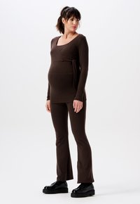 Braunes Umstandstop mit Wickeldesign und Bindedetail, kombiniert mit passenden ausgestellten Hosen. Beide aus weichem Stoff; das Model trägt schwarze Schuhe.