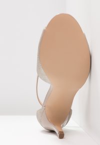 Chaussure beige à talons hauts positionnée semelle vers le haut contre un mur blanc, avec une tige texturée et un talon fin avec embout en caoutchouc.