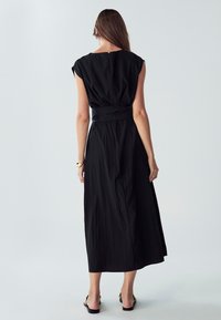 Črna dolga obleka brez rokavov z zategnjenim pasom, teksturirano tkanino in detajlom v obliki ključavnice na hrbtu. Ima tekoča silhueto in je dolga do gležnjev.