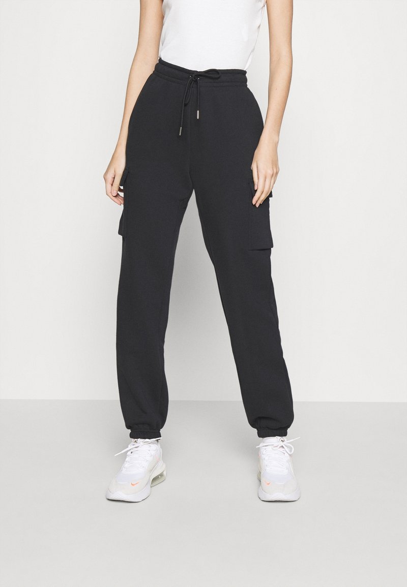 Černé joggingové kalhoty s páskem na stažení, bočními cargo kapsami a zúženými manžetami, vyrobené z měkkého a elastického materiálu. V kombinaci s bílými teniskami.