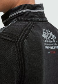 Schwarzer Pullover mit hohem Kragen, strukturiertem Stoff, kontrastierenden grauen Nähten und aufgedrucktem Schriftzug auf der Rückseite.