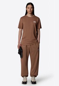 Hnedé bavlnené tričko a zodpovedajúce tepláky s malým logom na hrudi a boku, v kombinácii s čiernymi topánkami a šiltovkou.