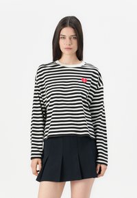 Donna con lunghi capelli scuri che indossa una maglietta a maniche lunghe a righe bianco e nera con un logo a forma di cuore rosso e una gonna plissettata nera.