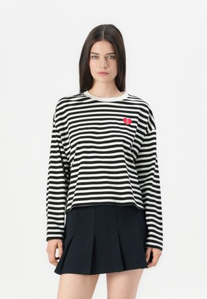 Femme avec de longs cheveux noirs portant un t-shirt rayé noir et blanc à manches longues arborant un logo en forme de cœur rouge et une jupe plissée noire.