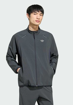Jonge man die een donkergrijs zip-up sportjack met Adidas-logo draagt, handen in de zakken, staand tegen een effen lichte achtergrond.