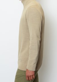 Homme portant un pull à col roulé beige et un pantalon olive, vu de profil avec le bras droit détendu le long du corps.