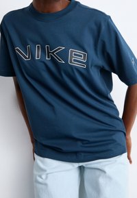 Osoba v modrém tričku Nike s velkými obrysy písmen a světle modrých kalhotách, stojící na jednobarevném bílém pozadí.