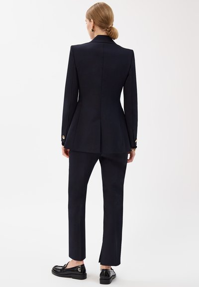 Femme aux cheveux blonds attachés, portant un blazer noir cintré, un pantalon slim assorti et des mocassins noirs, debout de dos.