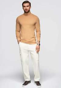 Beige Rollkragenpullover, taillierte Passform, kombiniert mit weißen Cargo-Hosen mit seitlichen Taschen. Braune Sneaker runden das Outfit ab.
