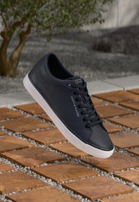 Baskets en cuir marine avec semelle en caoutchouc blanche, bout rond, lacets plats et détail de logo sur le côté. Positionnées au-dessus d'une surface en carrelage texturée.