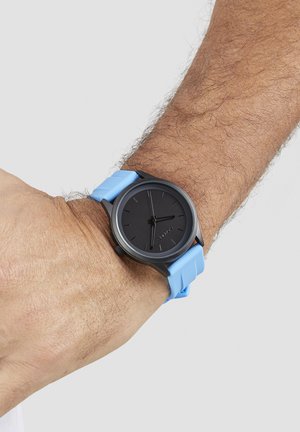 Orologio con quadrante nero, sottili indici delle ore, cassa grigia opaca e cinturino in silicone azzurro chiaro. Forma semplice e circolare con design minimalista.