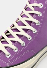 Purple canvas high-top sneaker with white laces, silver eyelets, and black rubber sole. Features contrasting stitching along the fabric.