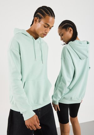 Mintgrønn hettegenser med frontlomme og ribbestrikkede mansjetter, båret av modeller. Stoffet ser mykt og teksturert ut.