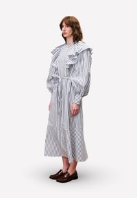 Frau steht in einem langen weißen Kleid mit vertikalen schwarzen Streifen, Rüschen an den Schultern, einer taillierten Taille und dunkelbraunen Slippers.