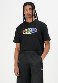 Black t-shirt featuring a multicolor "Adidas" logo. Short sleeves, loose fit, made of cotton fabric. Worn with black striped shorts.