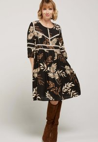 Femme portant une robe noire mi-longue avec un motif de feuilles beiges et des bottes en daim marron jusqu'au genou, se tenant devant un fond clair uni.