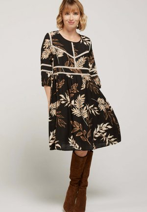 Femme portant une robe noire mi-longue avec un motif de feuilles beiges et des bottes en daim marron jusqu'au genou, se tenant devant un fond clair uni.