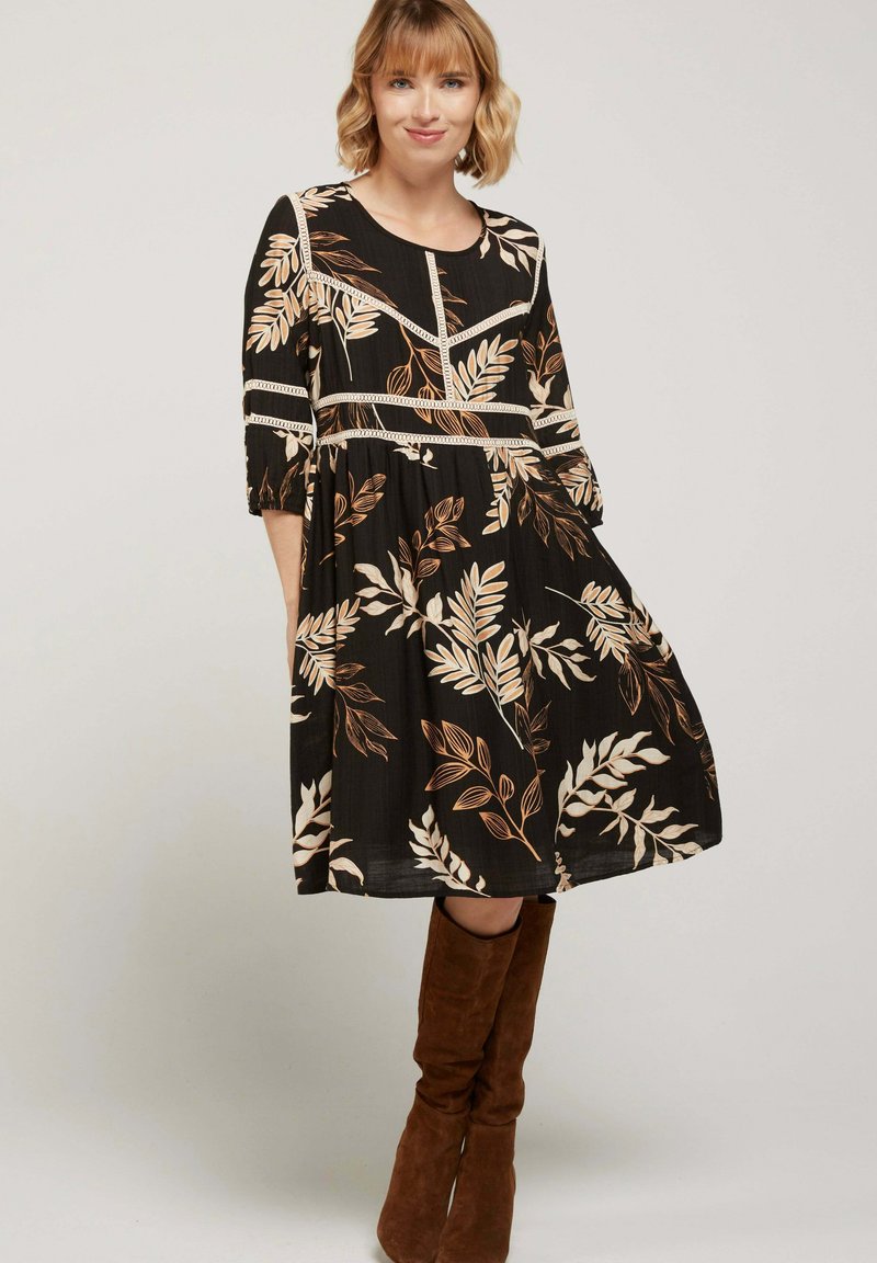 Femme portant une robe noire mi-longue avec un motif de feuilles beiges et des bottes en daim marron jusqu'au genou, se tenant devant un fond clair uni.