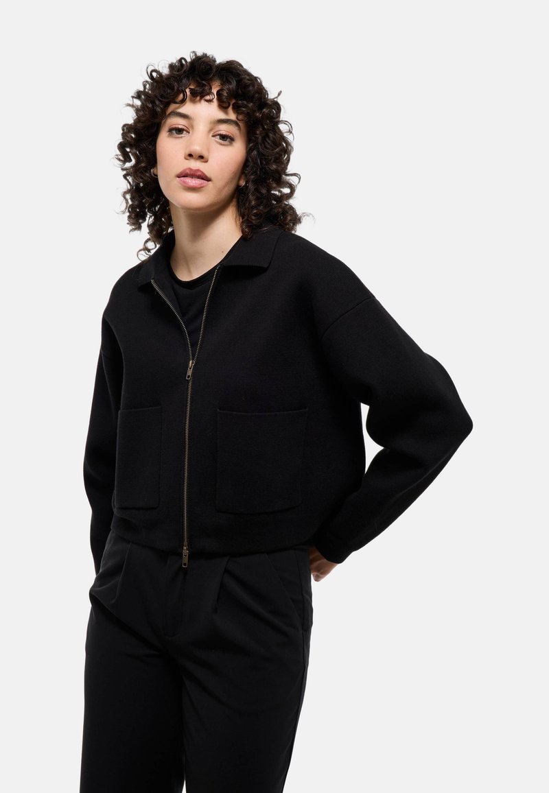 Veste noire courte avec une fermeture éclair à l'avant, deux grandes poches avant et un col cranté. Confectionnée dans un tissu texturé avec des manches longues.