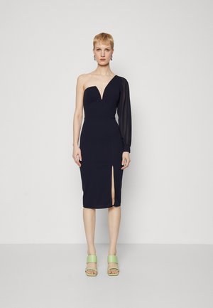 Robe ajustée bleu marine avec un décolleté en V profond, une manche longue transparente et une fente sur le côté ; associée à des sandales à plateforme vertes.