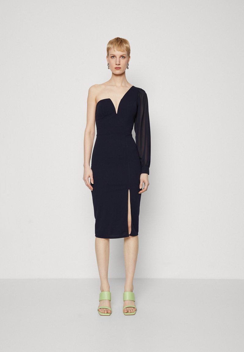 Robe ajustée bleu marine avec un décolleté en V profond, une manche longue transparente et une fente sur le côté ; associée à des sandales à plateforme vertes.
