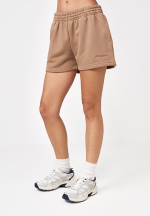 Kobieta ubrana w beżowe krótkie spodenki z elastycznym pasem, białe skarpetki i białe sportowe buty, stojąca na tle jednolitego białego tła.