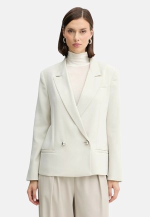 Blazer double boutonnage en tissu beige clair avec une texture lisse. Doté de boutons argentés et de deux poches latérales.