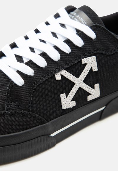 Baskets noires avec lacets blancs et logo fléché blanc brodé sur le côté, avec une semelle noire et un détail rayé blanc.