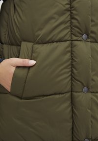 Vesta puf cu design matlasat de culoare verde, având o buzunar vizibil și capse negre, evidențiind o textură netedă și lucioasă.