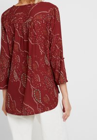 Blouse bordeaux avec des motifs floraux et paisley, manches trois-quarts, dos froncé et ourlet arrondi. Texture de tissu lisse.
