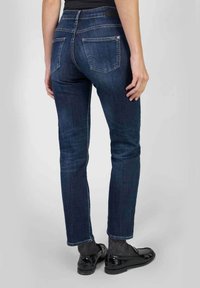Donkerblauwe, slim-fit jeans met een middelhoge taille, vijf zakken en subtiele stiksels, weergegeven vanaf de achterzijde met zwarte schoenen.