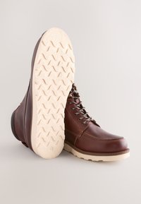 Hnedé kožené čižmy s hladkým povrchom, okrúhlou špičkou a kontrastnou bielou gumovou podrážkou so vzorom cik-cak. Kovové očká so šnúrkami.