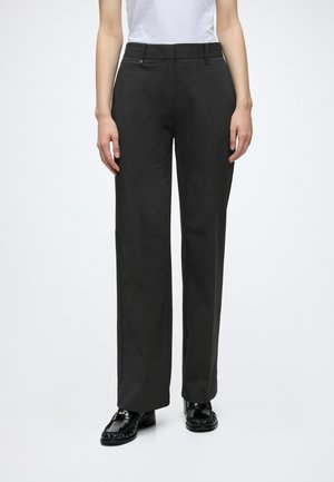Personne portant un pantalon noir à jambes droites, un haut blanc et des mocassins noirs, debout devant un fond clair uni.