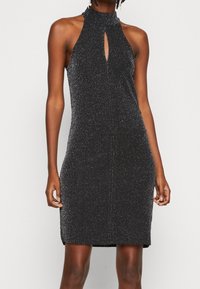 Svart ärmlös glittrig klänning med hög halsringning och detalj i form av nyckelhål framtill, bärs av stående person med avslappnade händer vid sidorna.