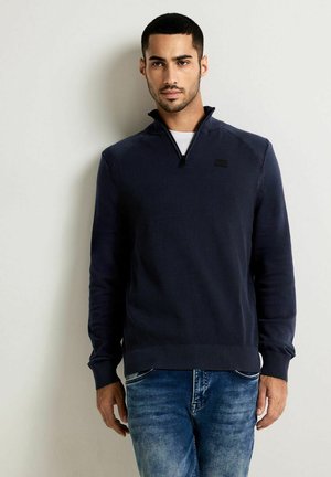 Marineblauer Zip-Pullover mit geripptem Muster, langen Ärmeln und kleinem Logo auf der Brust. Kombiniert mit hellblauen Jeans.